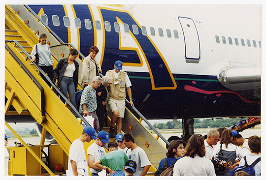 Light-skinned Kosovar refugees disembark off a plane using yellow stairs, while others stand nearby.