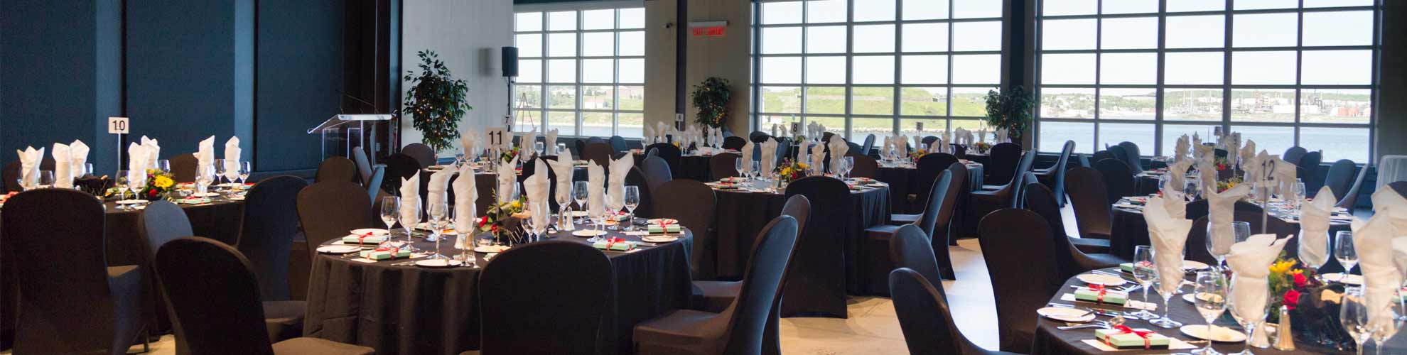 A room with floor to ceiling windows offering a view of the harbour and island and oval banquet tables and chairs set with dining wares.