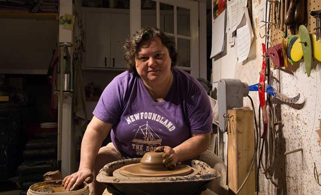 Woman in purple tee shirt sits in a workshop, throwing ceramic clay on a pottery wheel.