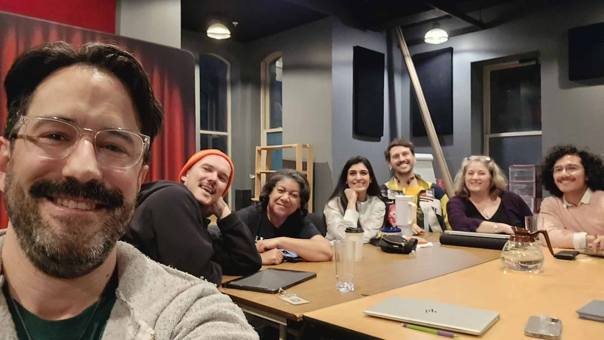 A selfie of the cast and crew sitting around a table with laptops.