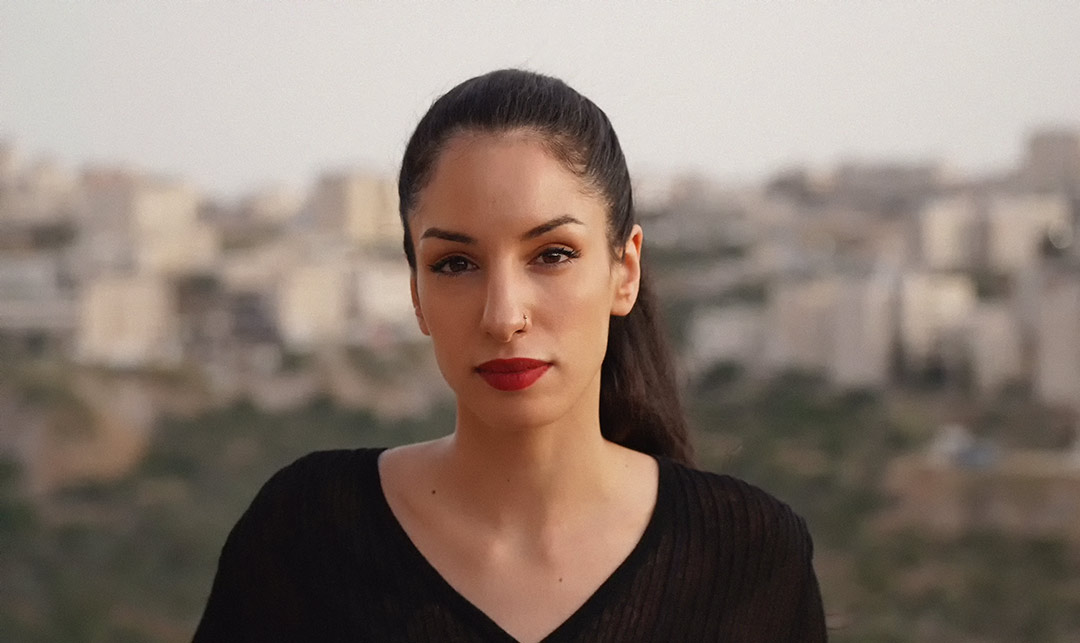 A portrait of Rana Nazzal Hamadeh against a blurred outdoor background, wearing a ponytail and red lipstick.