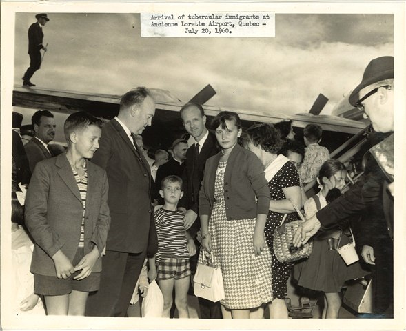 A sepia-toned photo of a large group of formally-dressed, light-skinned people on a tarmac with a plane behind them.