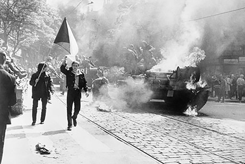 Black and white photo of people marching down a street, one person waves a flag, a tank in the background appears to be on fire.