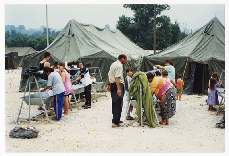 Men and women wash their hands in rows of metal basins, tents in the background.