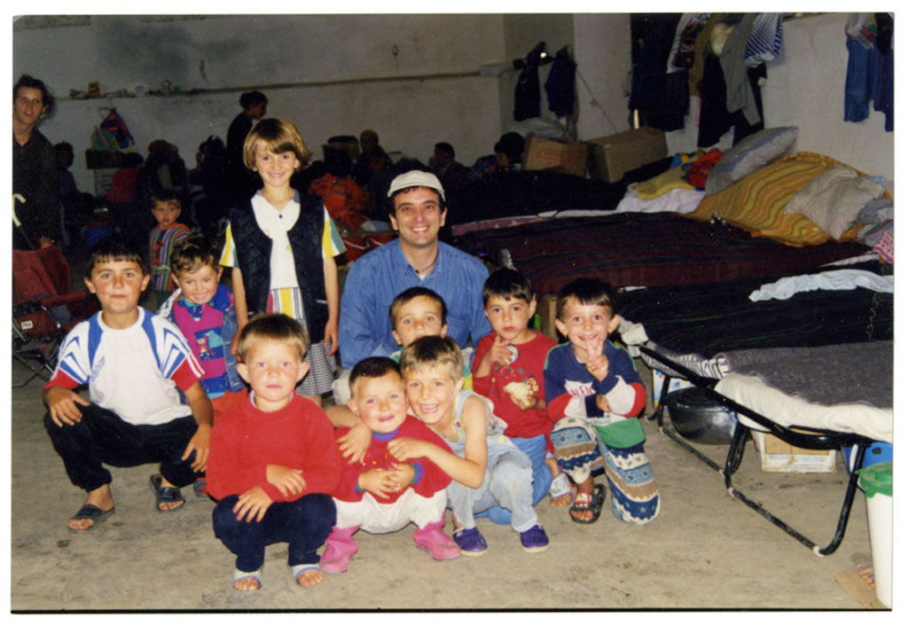 A man and a large group of children are smiling, kneeling next to a row of cots.
