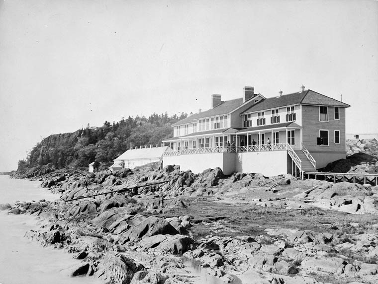 Black and white photograph of a large early 20th century wooden coastal building, a quarantine station, perched on rocky shores with a forested hillside behind it.