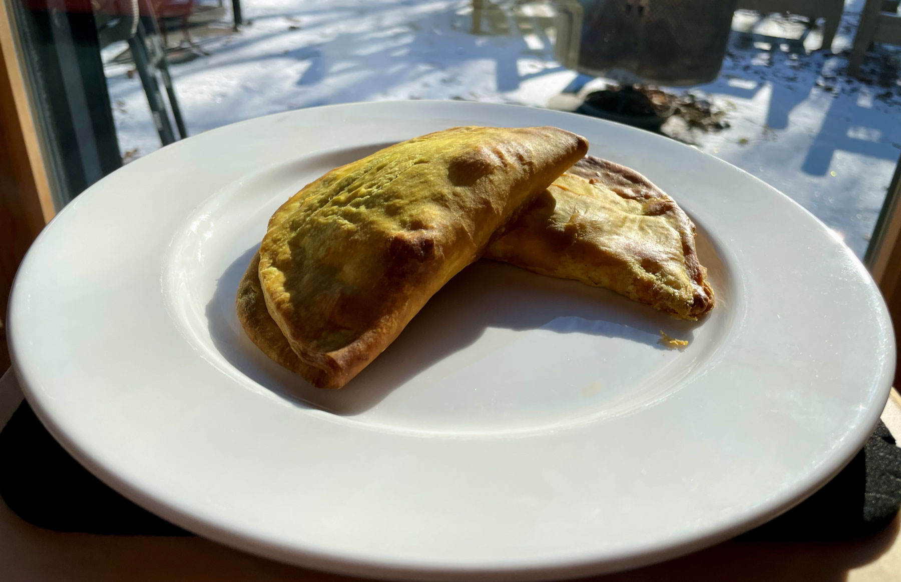 Two Jamaican patties on a white plate, behind the plate, in soft focus, through a window, a thin layer of snow covers the ground.