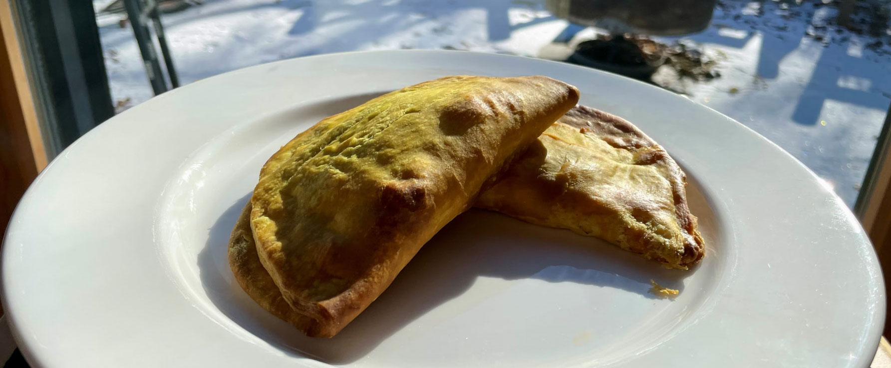 Two Jamaican patties on a white plate. Behind the plate, in soft focus, through a window, a thin layer of snow covers the ground.