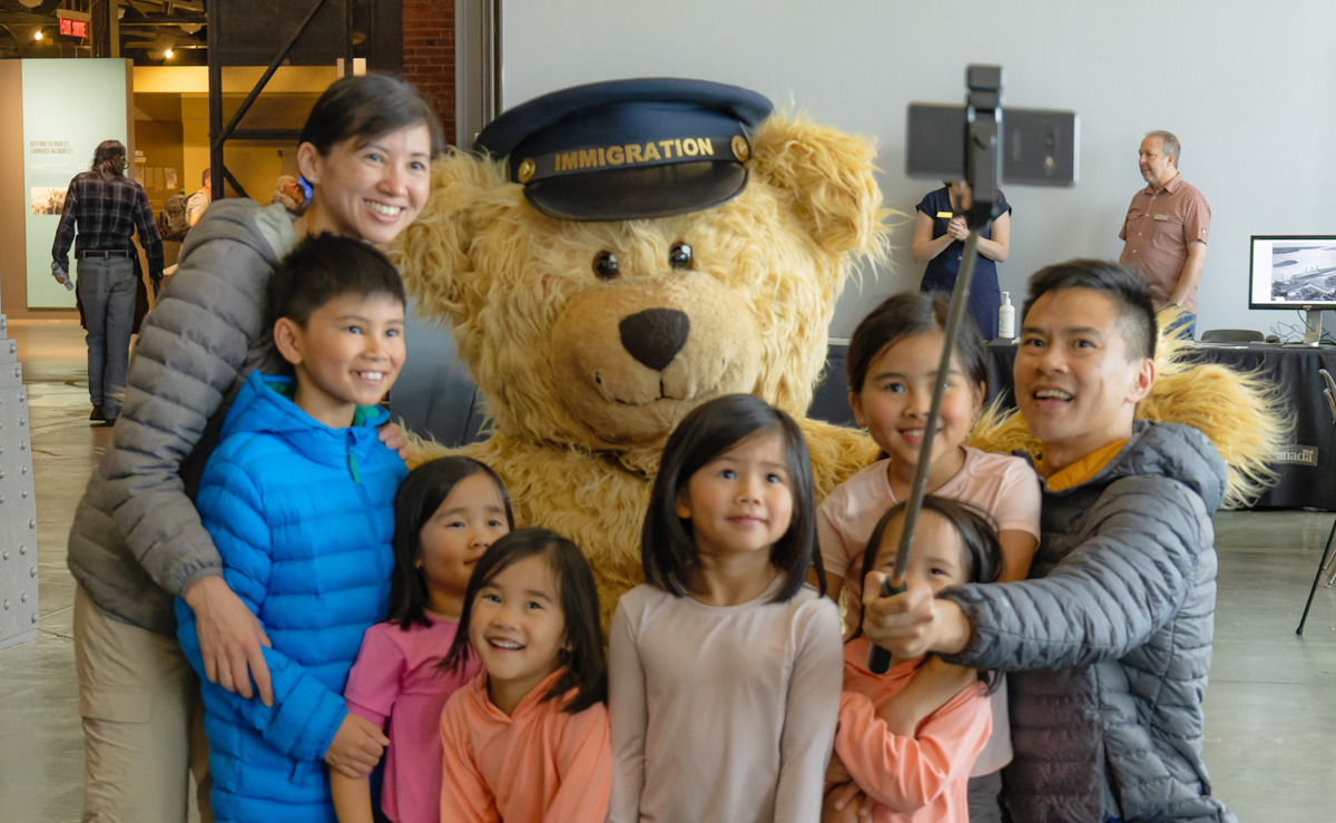 A large smiling bear mascot with a cap that says Immigration kneels, surrounded by six young children, a woman, and a man holding a selfie stick.