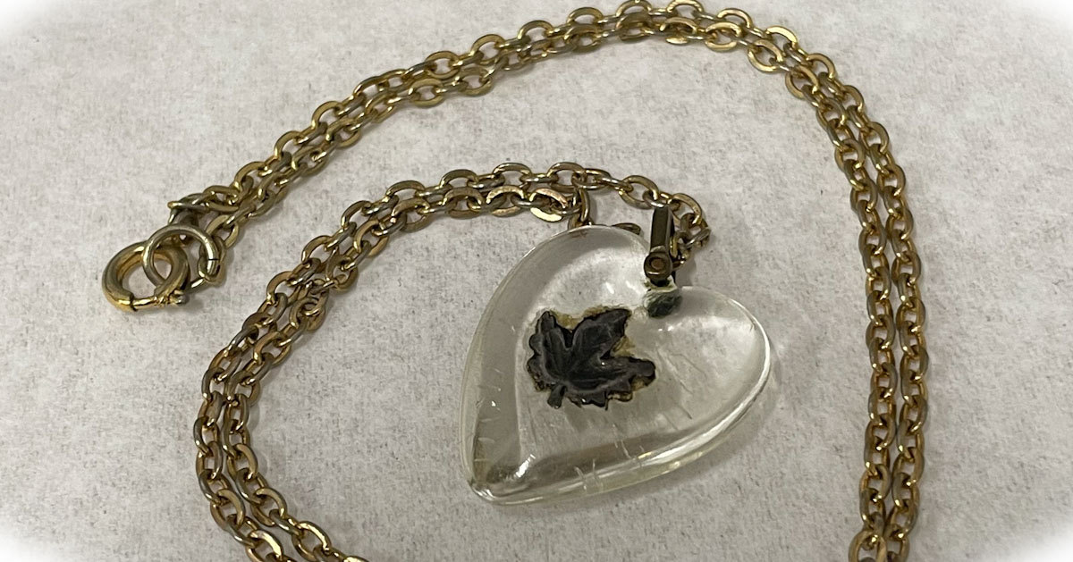 A glass pendant, a little bigger than a quarter, in the shape of a heart, with a small brass maple leaf on it, the chain of the necklace is gold.