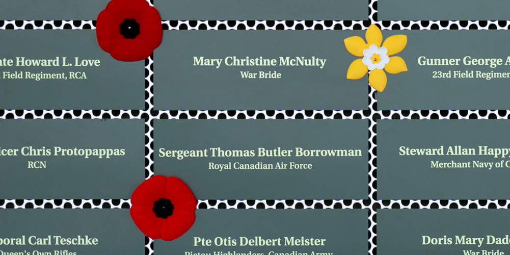 Green plaques inscribed with names of service people and war brides. Poppies and daffodil pins have been placed next to some plaques.