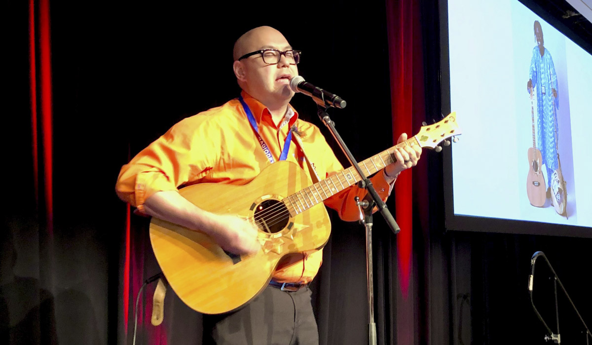 A middle-aged Japanese Canadian man plays a guitar on a temporary stage.