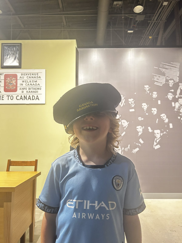 A boy, age 9, grins in an oversized Canada Immigration cap.