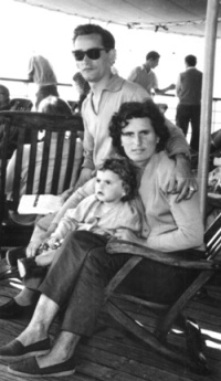 Woman seated on deck chair, with baby in lap and man on chair arm