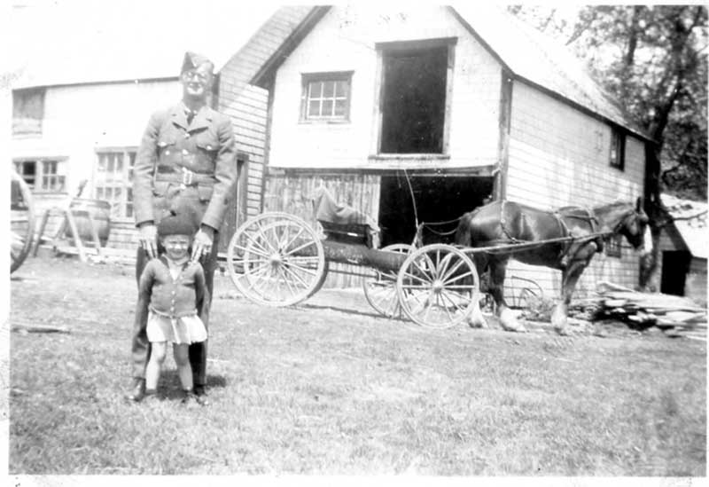 Uniformed man standing with small girl in front of a house,a horse cart is behind them.