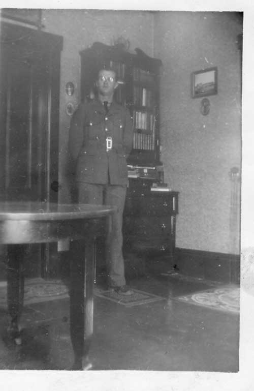 Uniformed man standing in a corner of a room in front of a book shelf behind a table.