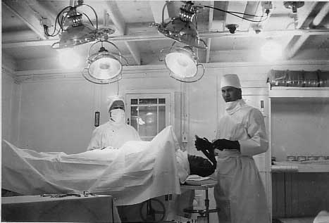 A man is lying on an operating table and two doctors wearing white gowns, caps and masks are standing beside him.