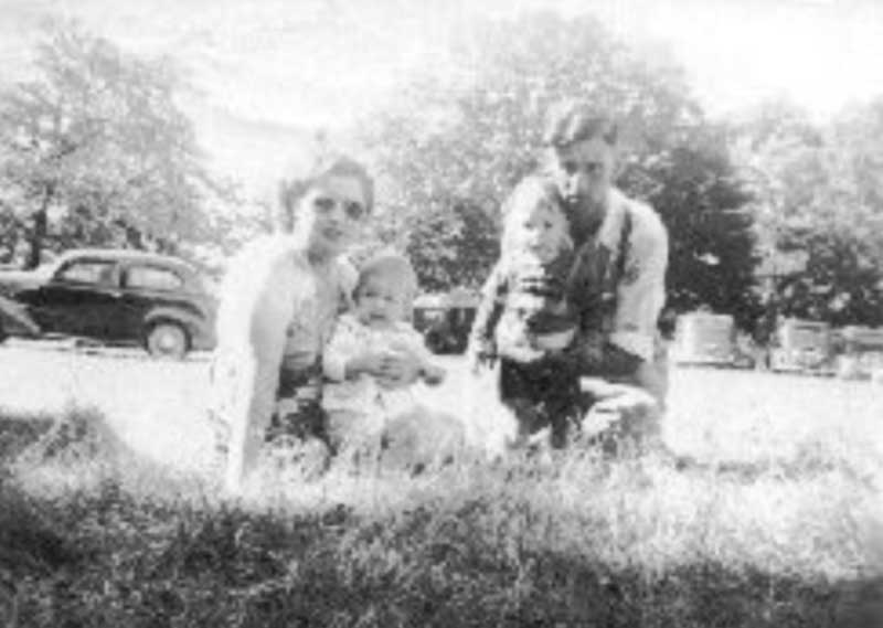A young man and woman sit on the grass, holding their children.