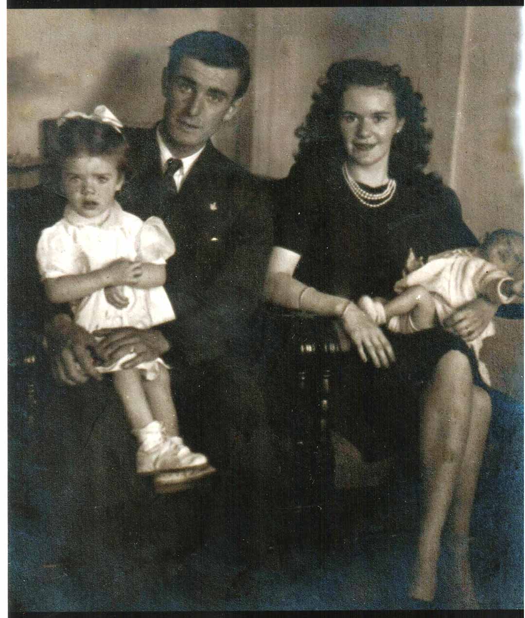 Les and Joan in a black and white photograph in the center of a sofa, Joan has her daughter Rosalind on her lap and Les has his daughter Pauline on his.