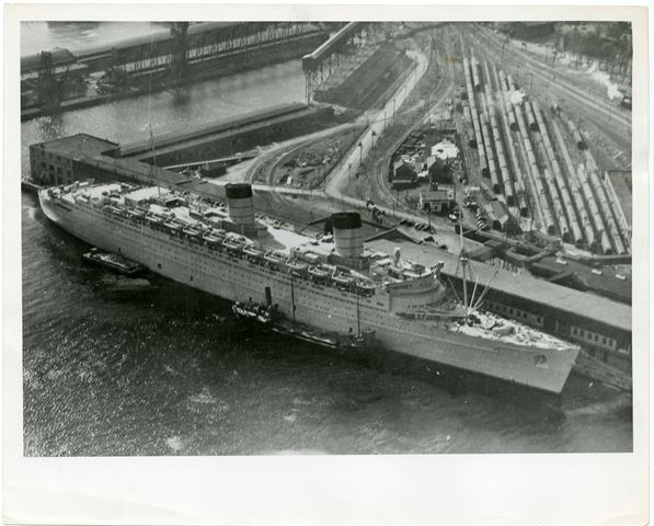 Viewed from the air, a large troopship painted wartime grey docks at a series of low waterfront sheds, with trainyards and a second shed building behind. Tugboats assist the ship in docking. To the left of the image and in the background, are some of the grain and cargo structures for nearby piers.