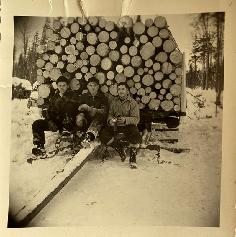 Three young men sit in front of a huge pile of logs.