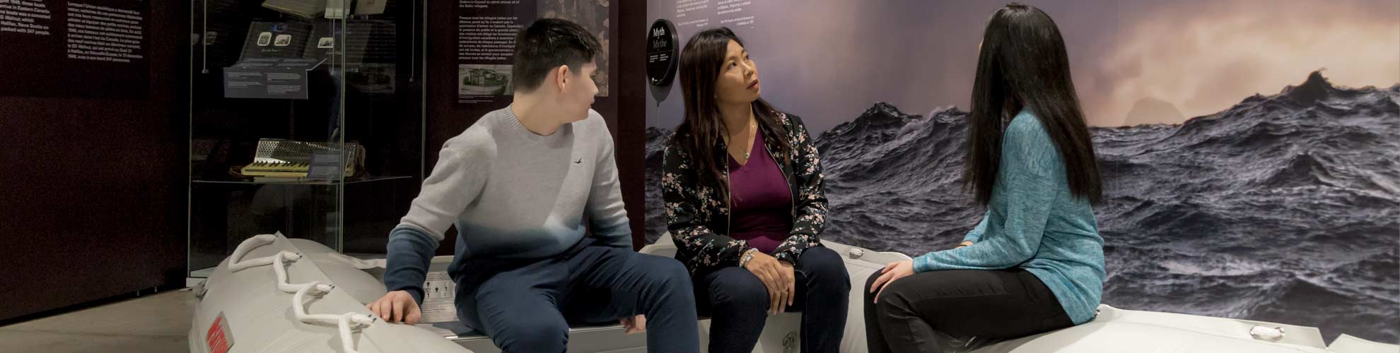 Three people sit in a lifeboat, looking at an exhibition panel of waves in the water.