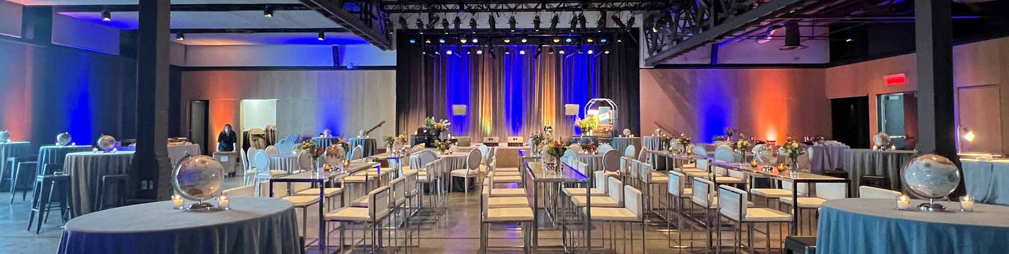 Tall clear bar tables and cruiser tables with blue linens have white bar stools for seating. The tables are decorated with blue linens, globe centerpiece and bud vases facing a stage with blue up lights.