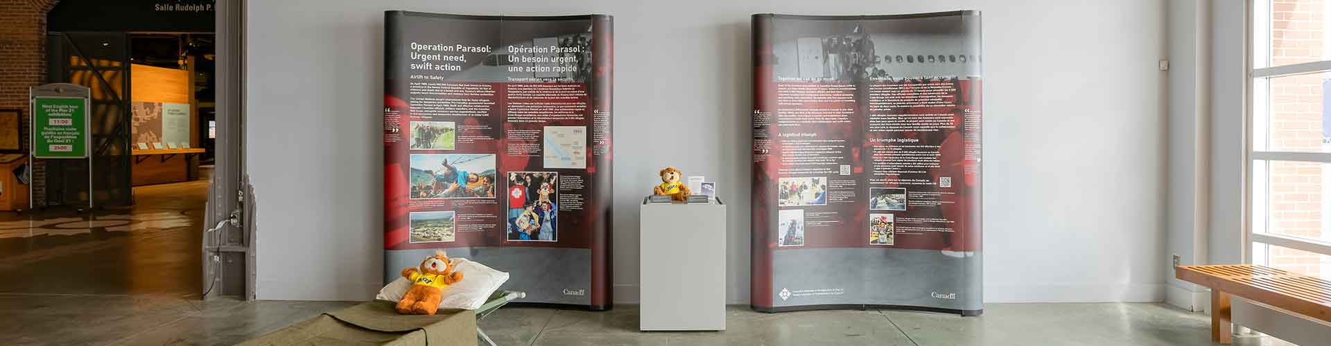 Two large stands with exhibition text and images. Two Zeddy bears are featured: one on a cot and one on a stand.