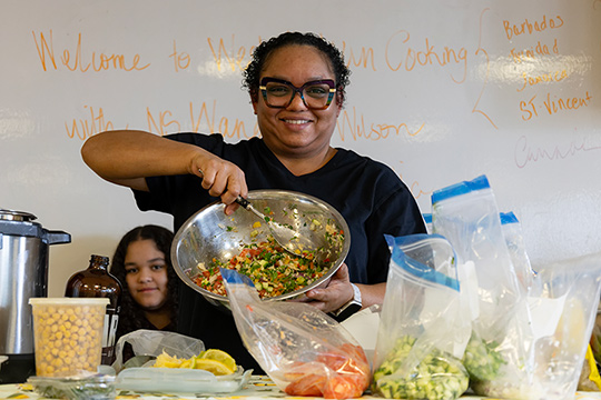 A smiling Caribbean woman holds up a metal bowl full of chopped vegetables.