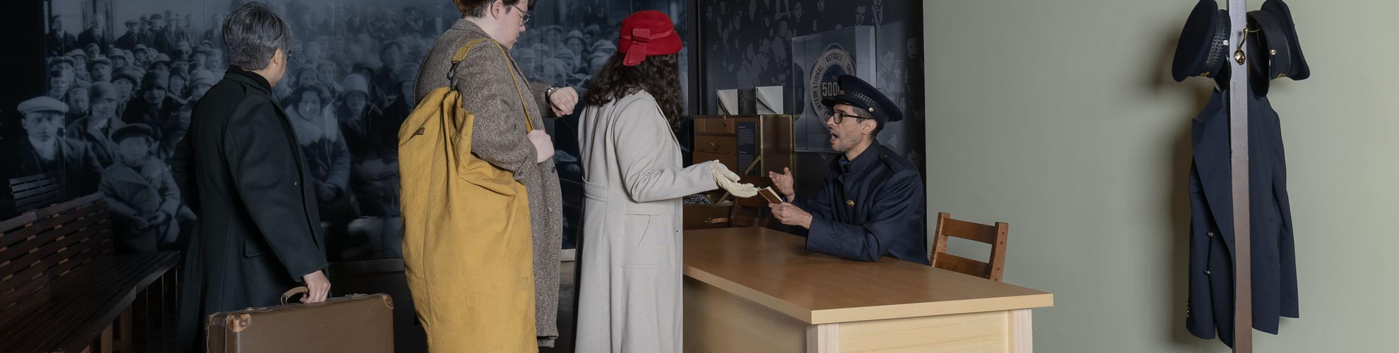 An immigration simulation showing a man in uniform at a desk speaking with three people lined up carrying old suitcases.