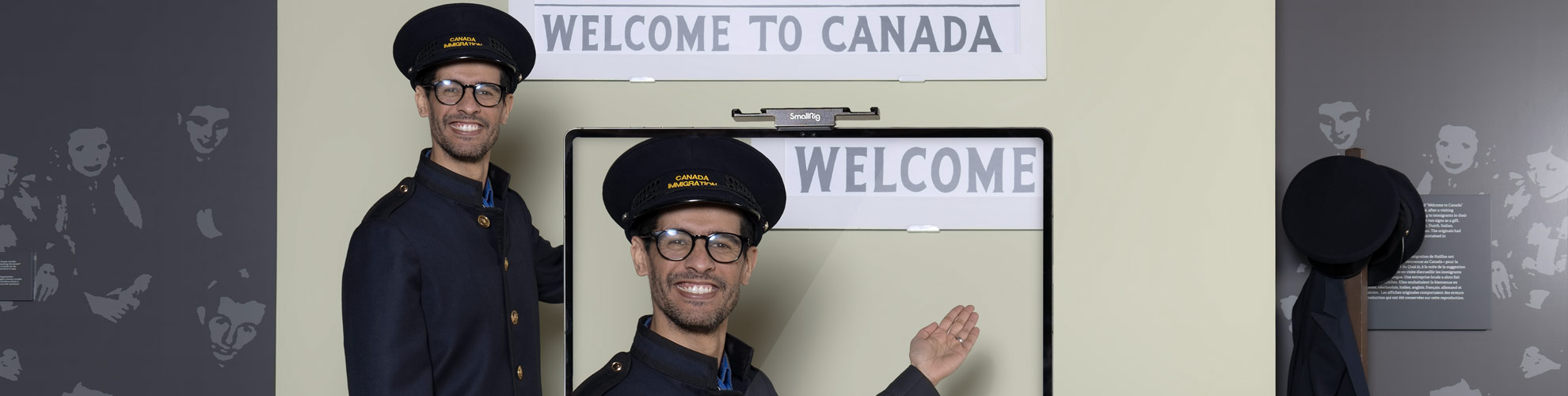 A smiling, uniformed man with medium skin tone gestures at a Welcome to Canada sign, standing in front of a camera.