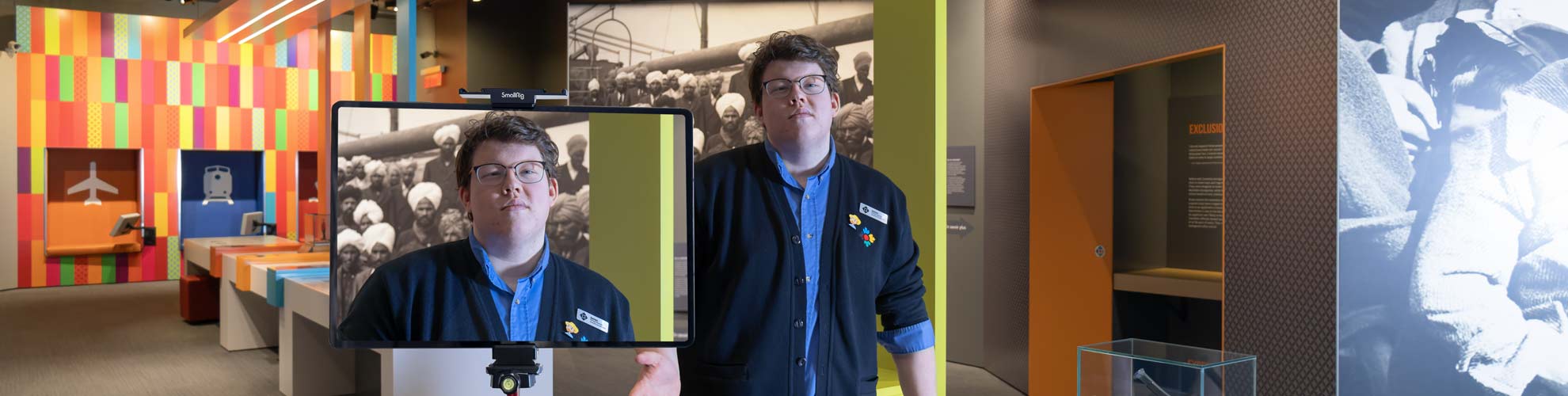 A white man in a Museum uniform stands in an exhibition and smiles into a large camera, the screen visible.