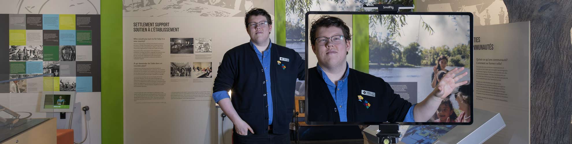 A white man in a Museum uniform stands in an exhibition and gestures into a large camera, the screen visible.