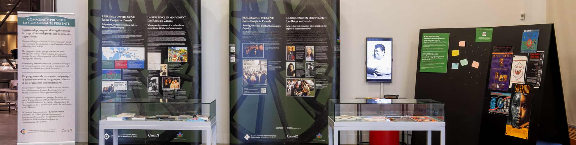 Roma People in Canada exhibit with informational panels, two glass display cases, and a black board with posters and notes.