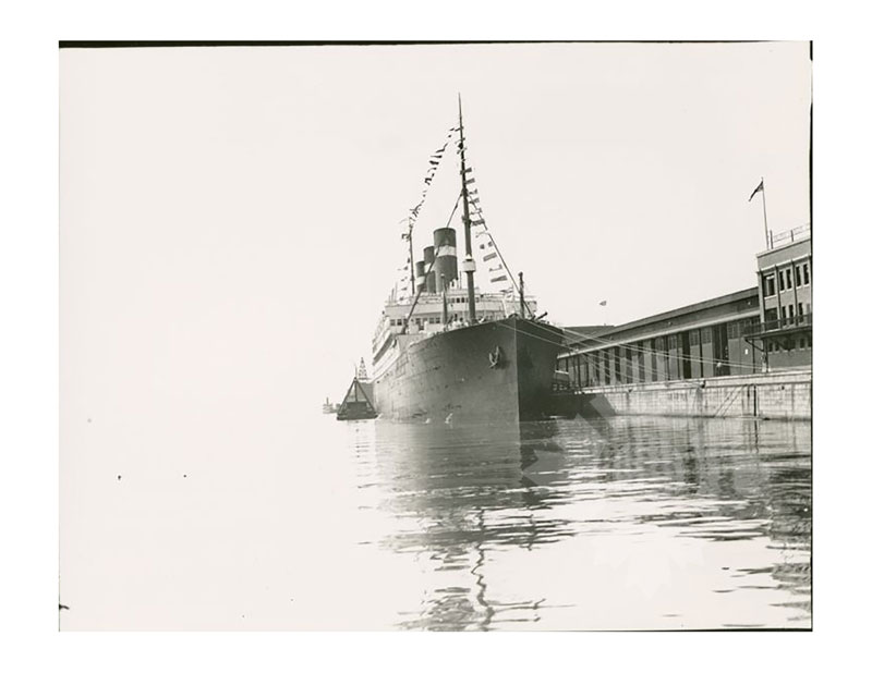Black and white image of SS Belgenland docked at a pier, seen from the bow, with a port building visible to the right.