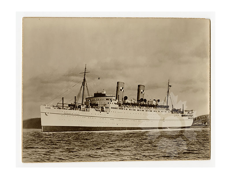 Black and white image of RMS Empress of Canada II at sea, broadside from port, with twin funnels and coastal hills behind.