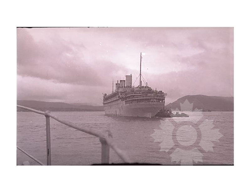 Black and white image of SS Île de France in harbour, seen from the bow, tugboat alongside and coastal hills behind.