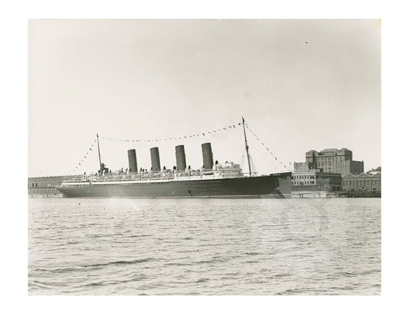 Black and white image of RMS Mauretania I docked in harbour, broadside from port, with four funnels and festive bunting.