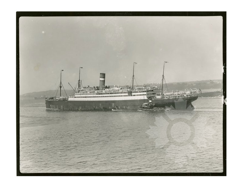 Black and white image of SS Nieuw Amsterdam I at sea, broadside from port, with a tugboat alongside.