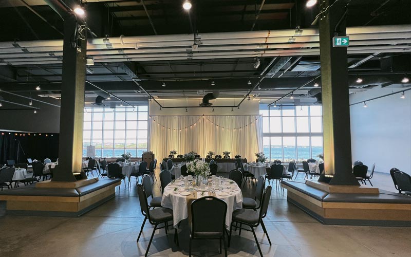 Wedding banquet set with oval banquet tables and a head table between two floors to ceiling windows overlooking the harbour. Behind the head table, the wall is decorated with white drapes and lights.