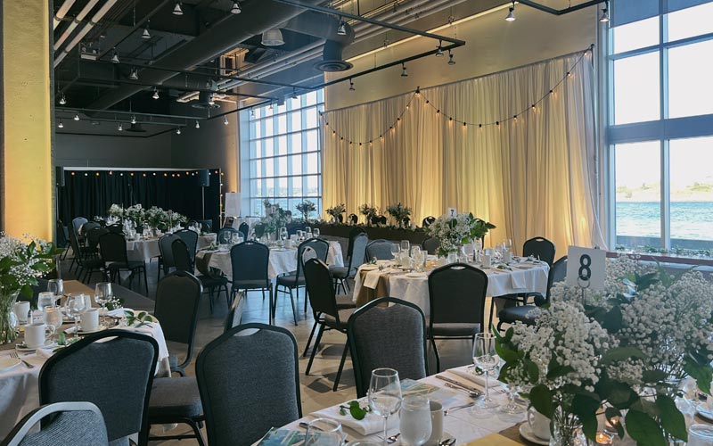 Wedding banquet set with decorated oval guest tables and a long head table, a small stage for a band, and large floor to ceiling windows.