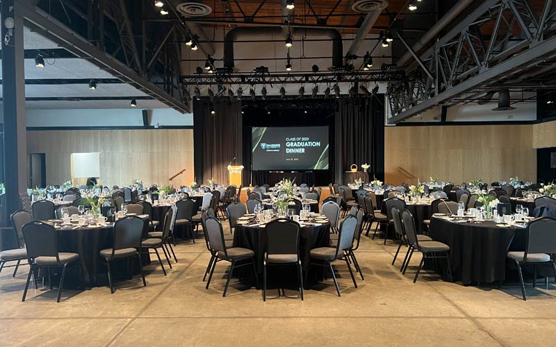 The venue is set for an awards banquet with oval tables, banquet chairs, and centerpieces on each table. A built-in stage is visible and contains a podium, projection screen and an awards table.