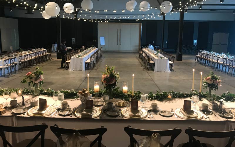 The room is set for a wedding with a long head table on a stage, decorated with garlands, candles and bouquets. Dining tables are set in four long rows with wooden chairs on both sides.