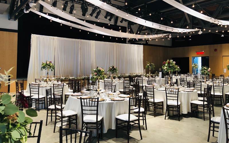 The venue is set for a wedding, including string lights and white drapes across the ceiling, a backdrop behind the head table, and guest oval tables.