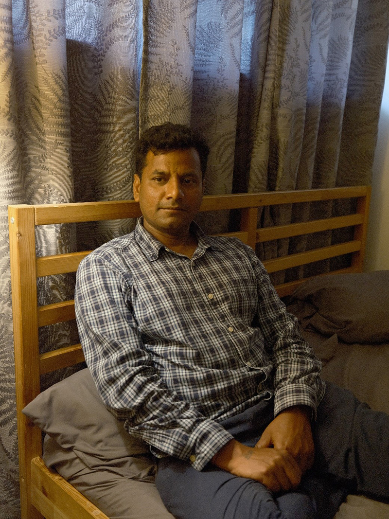 A man is sitting on a wooden bed with curtains behind it.
