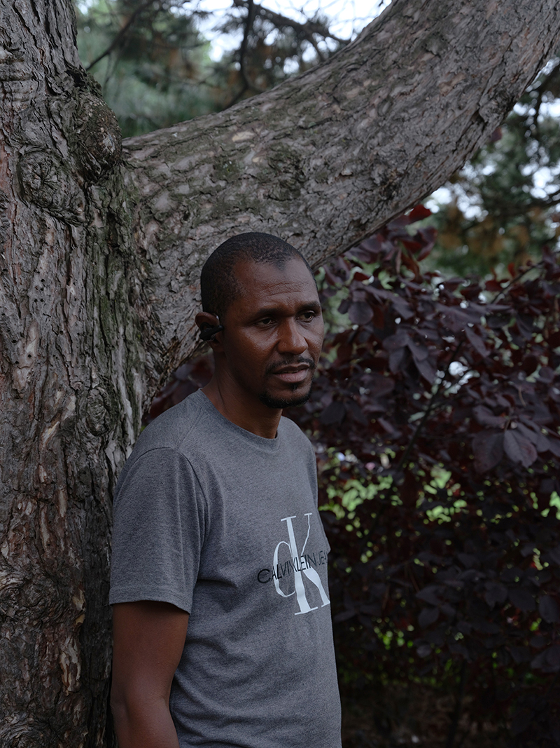A man in a grey tee shirt leans against a big tree.