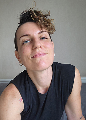 A portrait of Annie Valentina smiling at the camera, wearing a black tank top and buzzed hair with a curly bang.