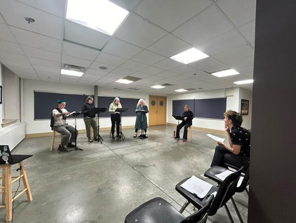 Five people stand or sit in a row at podiums while Annie sits and watches, chin rested on her hand.