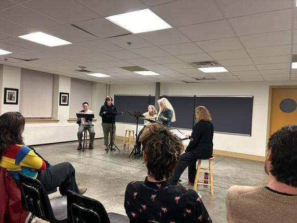 Five people stand or sit in a row at podiums while three people watch in the audience.