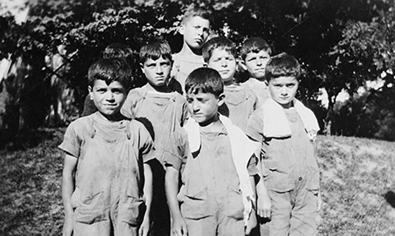 A group of seven young boys wearing overalls standing in a field with trees behind them.